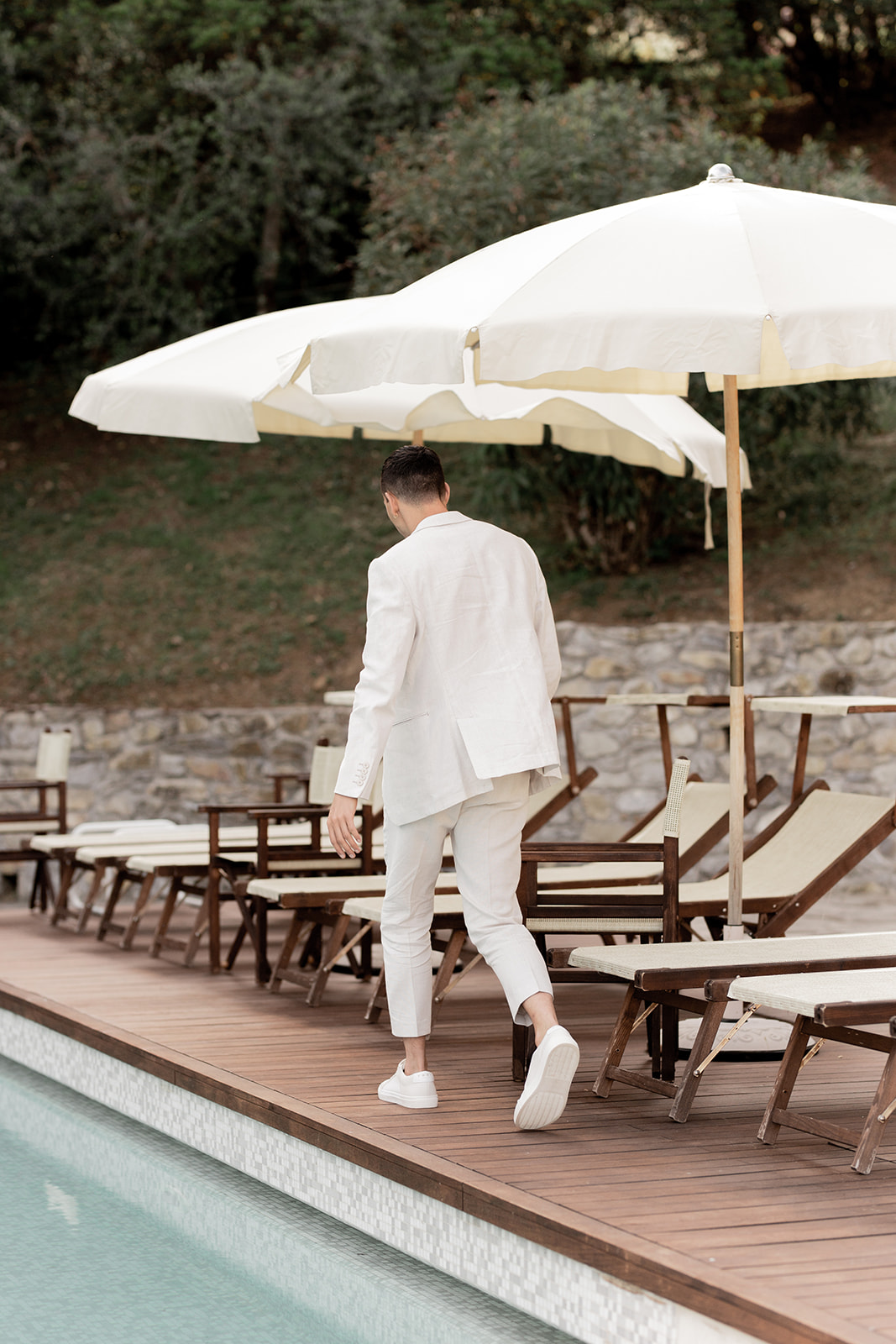 A person in a white outfit walks by a pool under large white umbrellas on a wooden deck, surrounded by lounge chairs and greenery in the background.