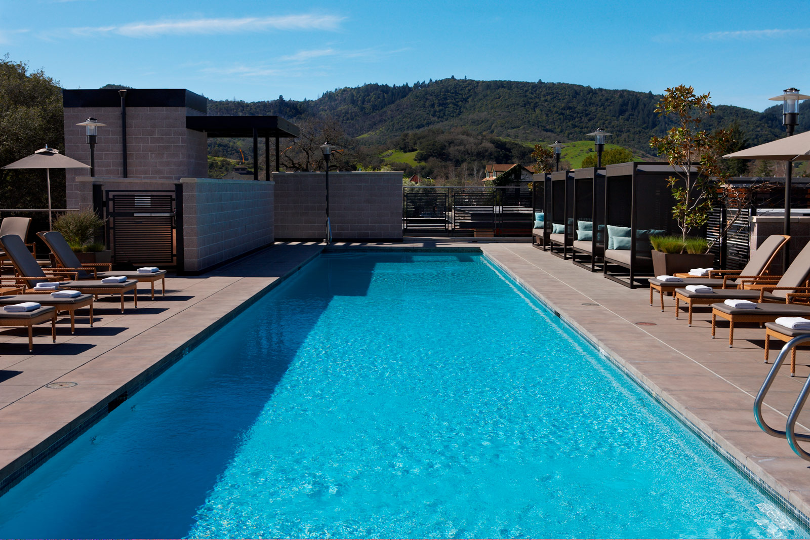 A long rectangular outdoor pool with lounge chairs and sun umbrellas around it. Trees and a hilly landscape are visible in the background under a clear blue sky.