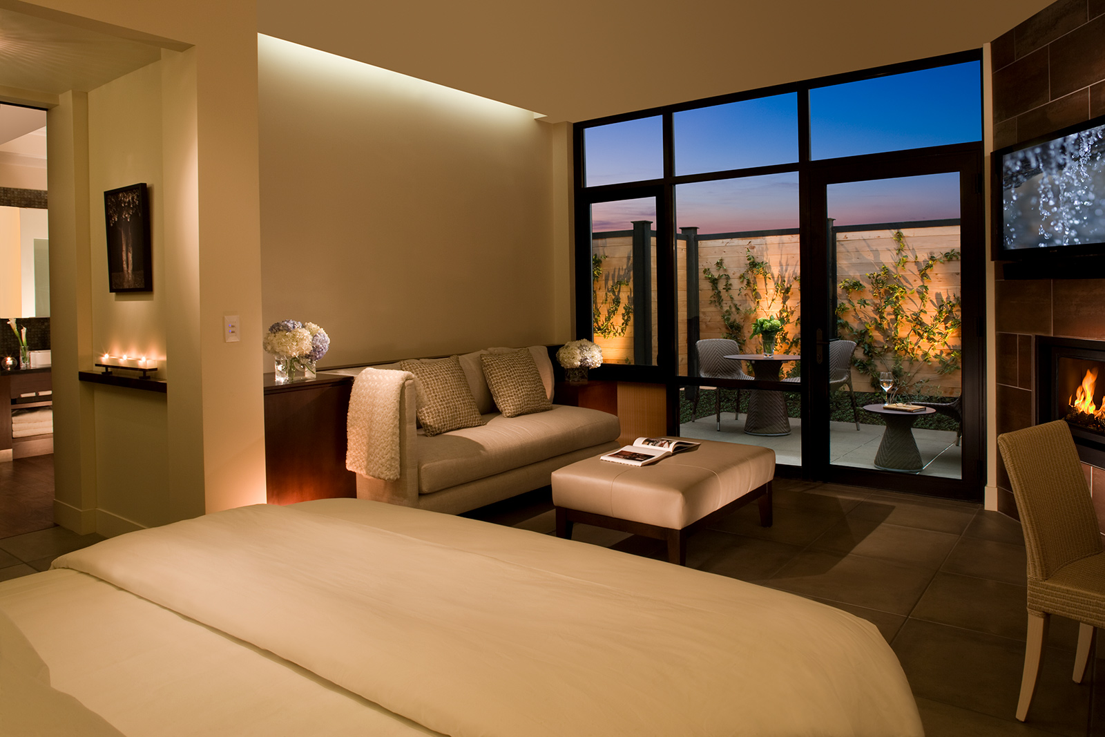 A modern bedroom features a bed, sofa, and a wall-mounted TV. Large glass doors open to an outdoor patio with seating and plants. Candles and flowers add decorative touches inside.