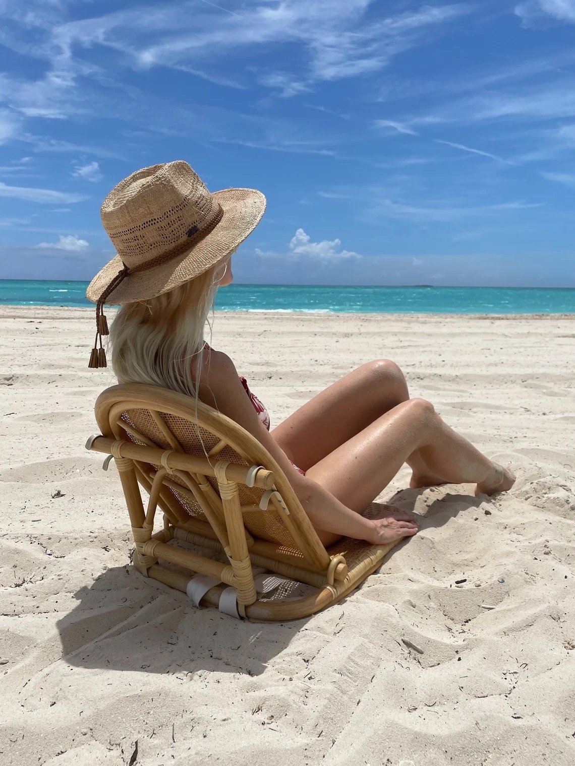 A person wearing a sun hat and swimsuit sits on a reclining beach chair facing the ocean on a sandy beach under a bright blue sky.