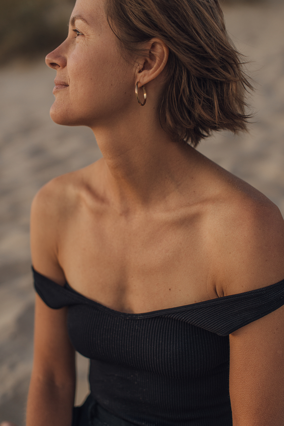 A woman with short hair wearing a black off-the-shoulder top and gold hoop earrings looks to the side while sitting outdoors.