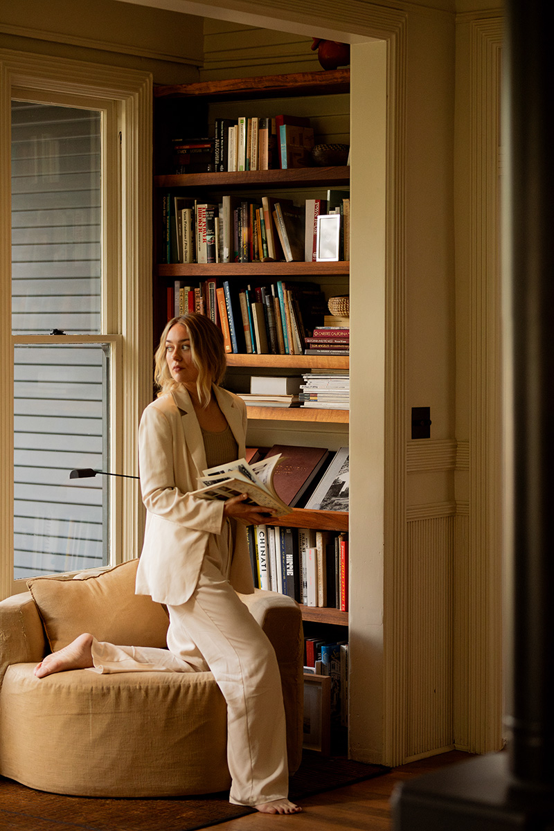 A person dressed in light-colored clothing sits on a beige armchair next to a bookshelf, holding an open book in front of a window.