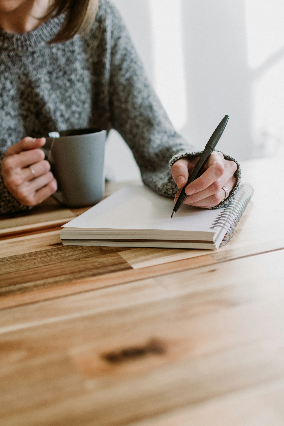 A person is writing in a spiral notebook while holding a mug at a wooden table.