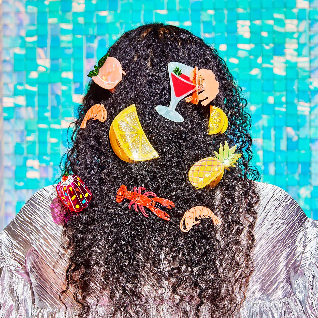 Person with curly hair adorned with colorful clips shaped like food and drinks, including lemon slices, shrimp, and a cocktail, against a shiny blue tile background.