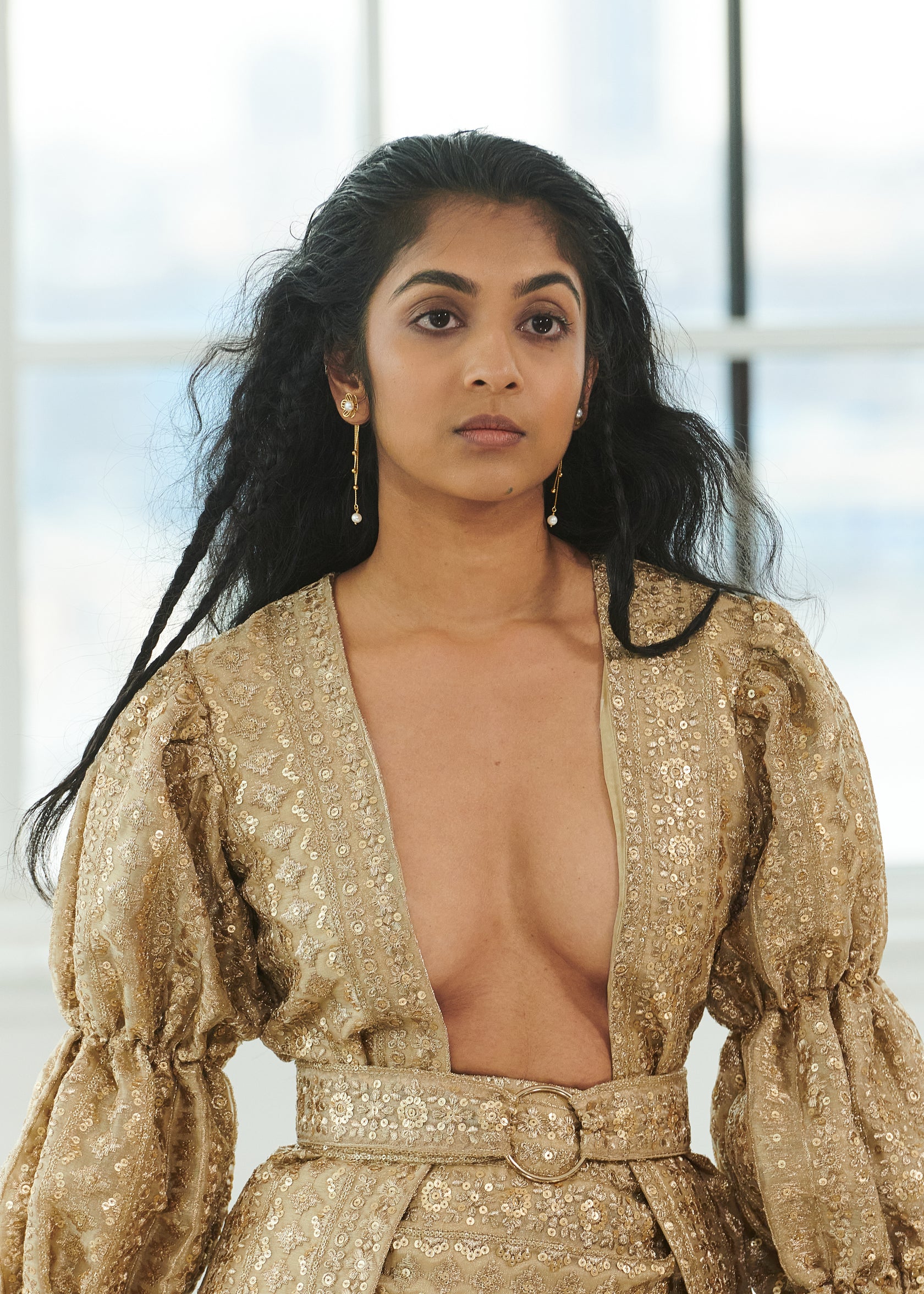 Person wearing a gold, patterned dress with long sleeves and deep neckline, standing indoors in front of a window.