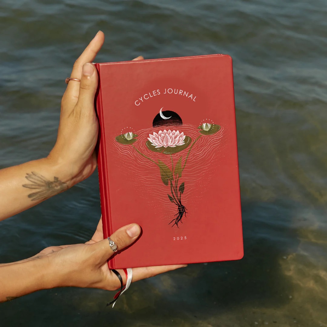 Hands holding a red journal titled "Cycles Journal 2025" with a design of a flower, crescent moon, and eyes, set against a backdrop of water.