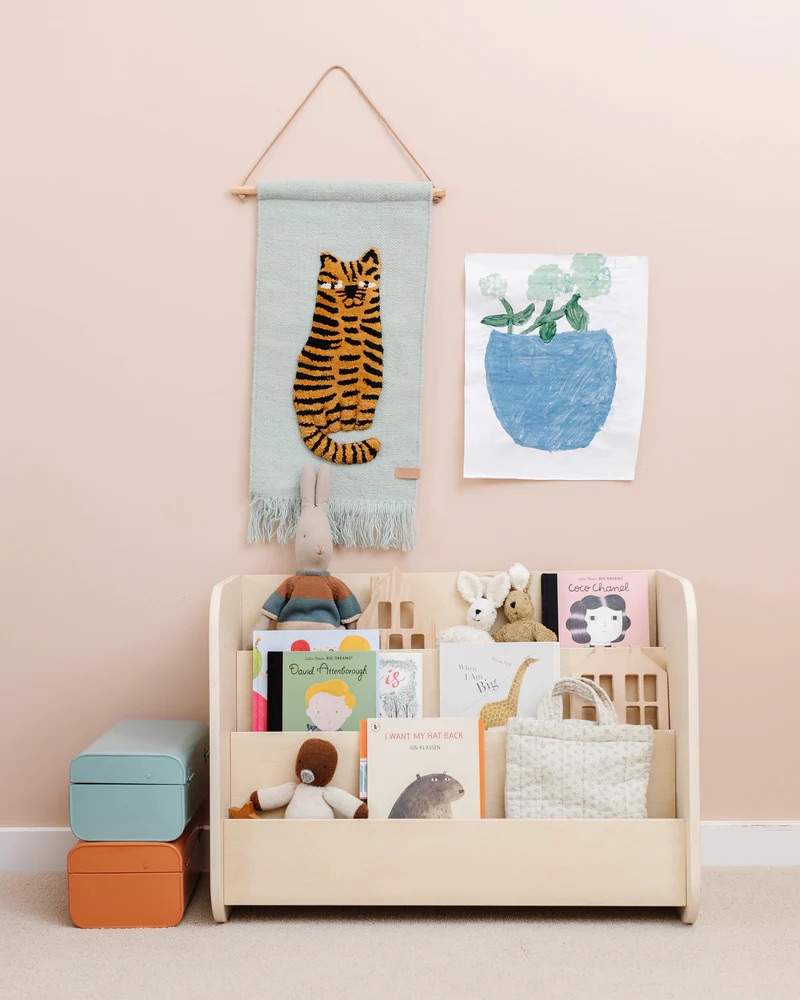 Children's play area with a wooden shelf holding books, toys, and storage boxes. A tiger wall hanging and a plant drawing are on the peach wall.