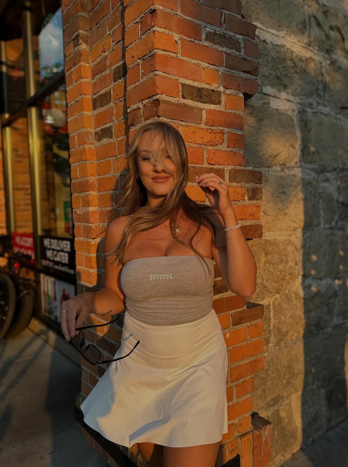 A woman stands against a brick wall, wearing a gray tube top and white skirt. Her hair is blowing across her face, and she is holding sunglasses in one hand.