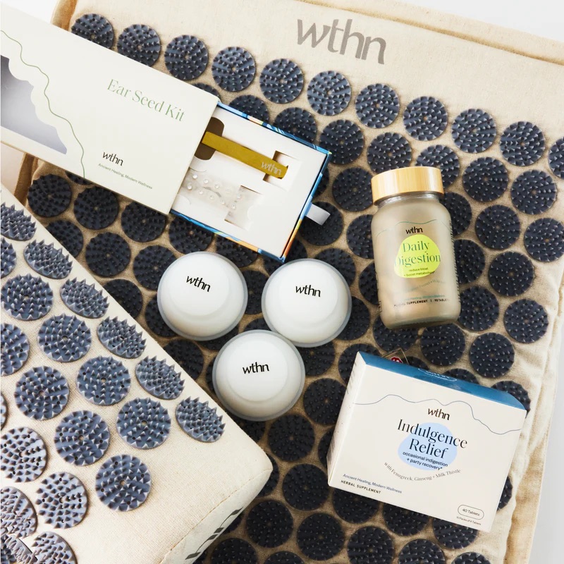 A collection of wellness products including an ear seed kit, supplements, and three small jars, displayed on a textured mat.