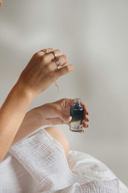 Person holding a dark liquid in a small, clear dropper bottle, with a dropper raised above. The person is wearing several rings.