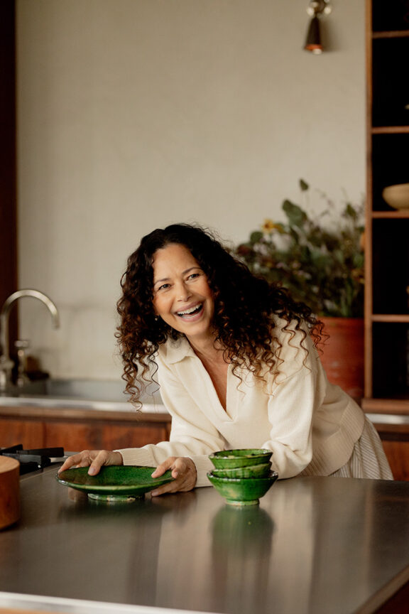 A woman with curly hair smiles while leaning on a kitchen counter, holding a green plate near stacked green bowls. There's a sink and a potted plant in the background.