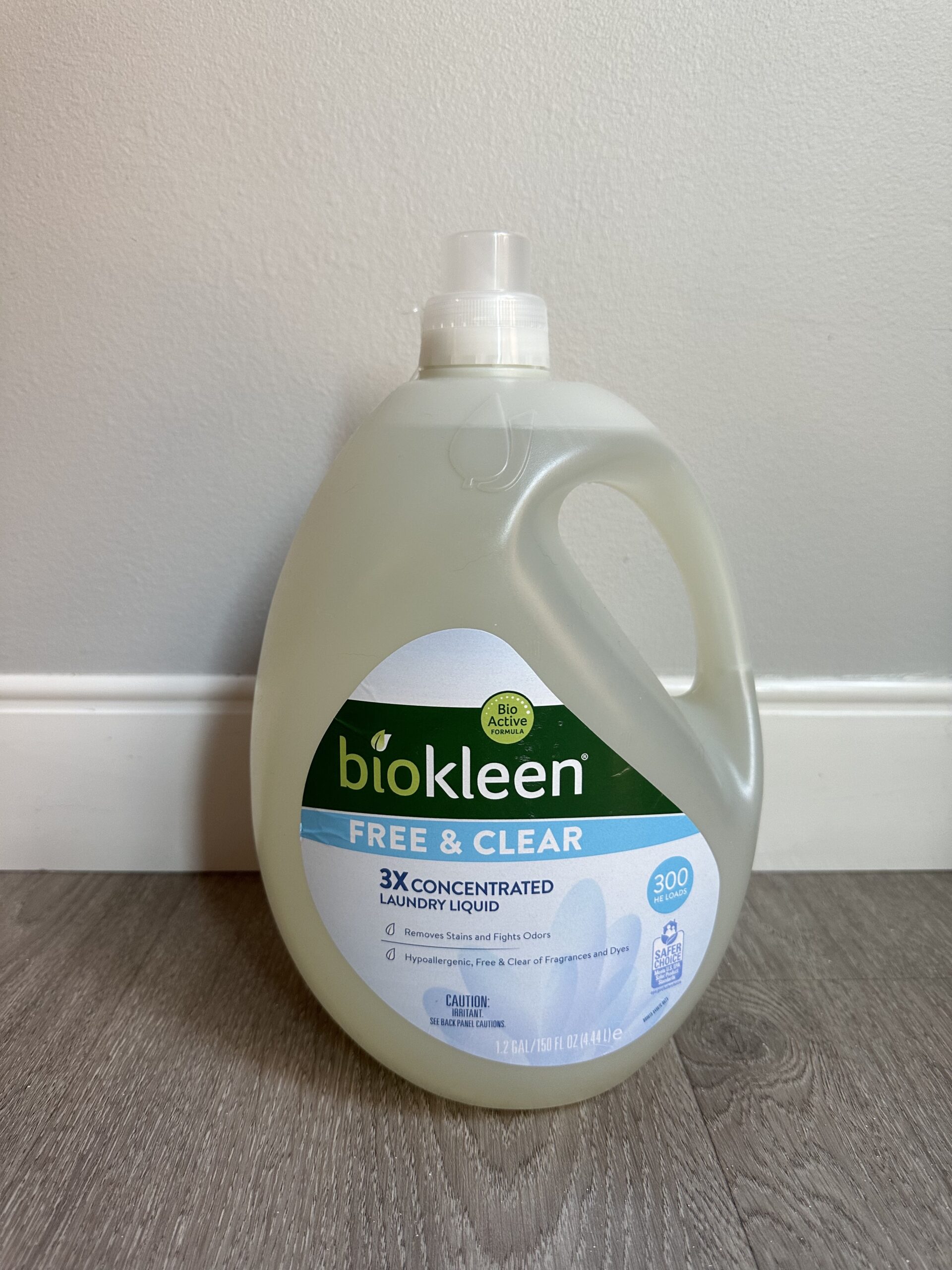 A bottle of Biokleen Free & Clear 3X Concentrated Laundry Liquid is placed on a wooden floor against a white baseboard. The bottle is clear and has a capacity of 150 ounces.