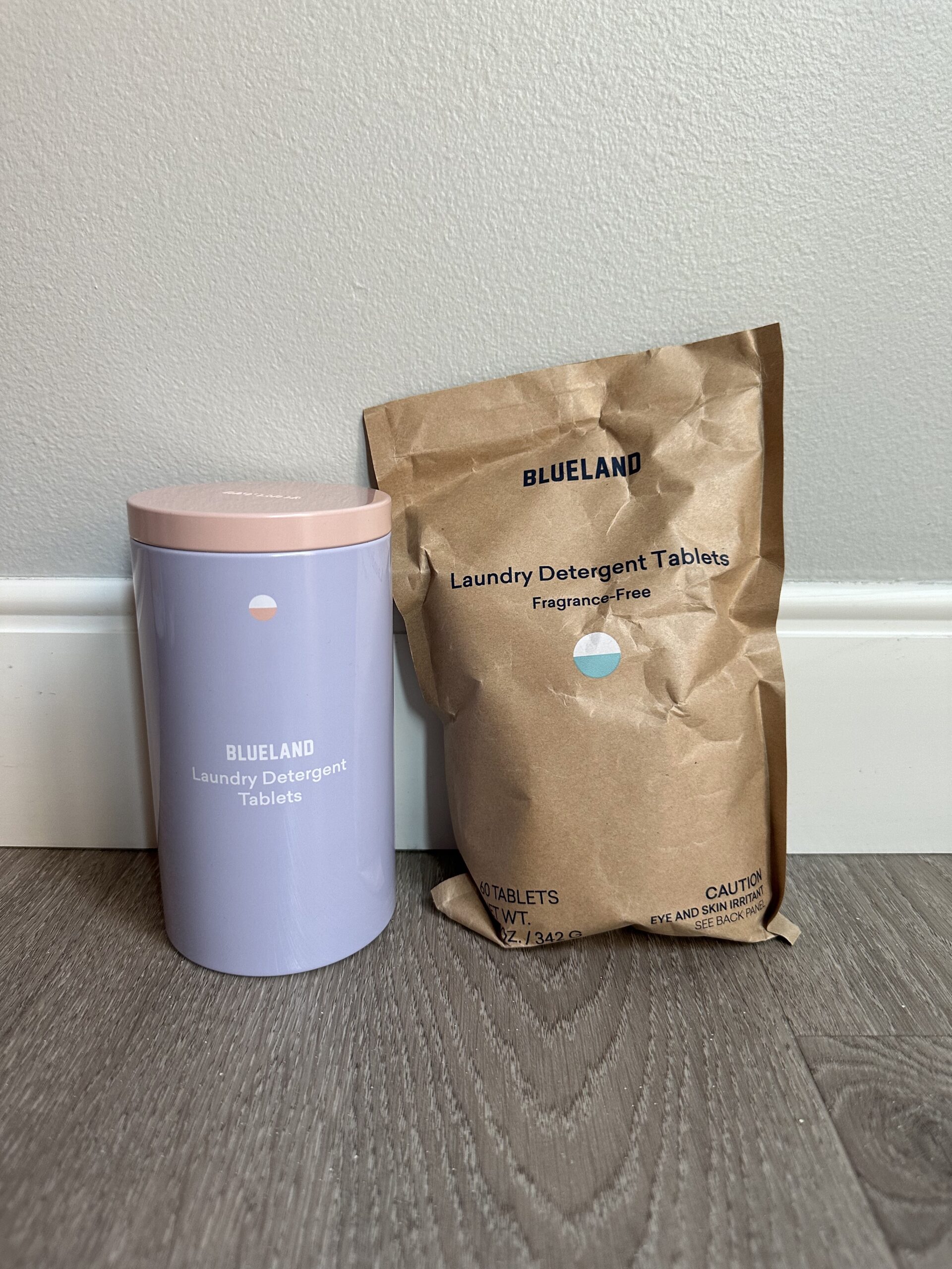 Blueland laundry detergent tablets in a cylindrical container and a brown paper refill bag, both labeled "fragrance-free," placed on a wooden floor against a light gray wall.