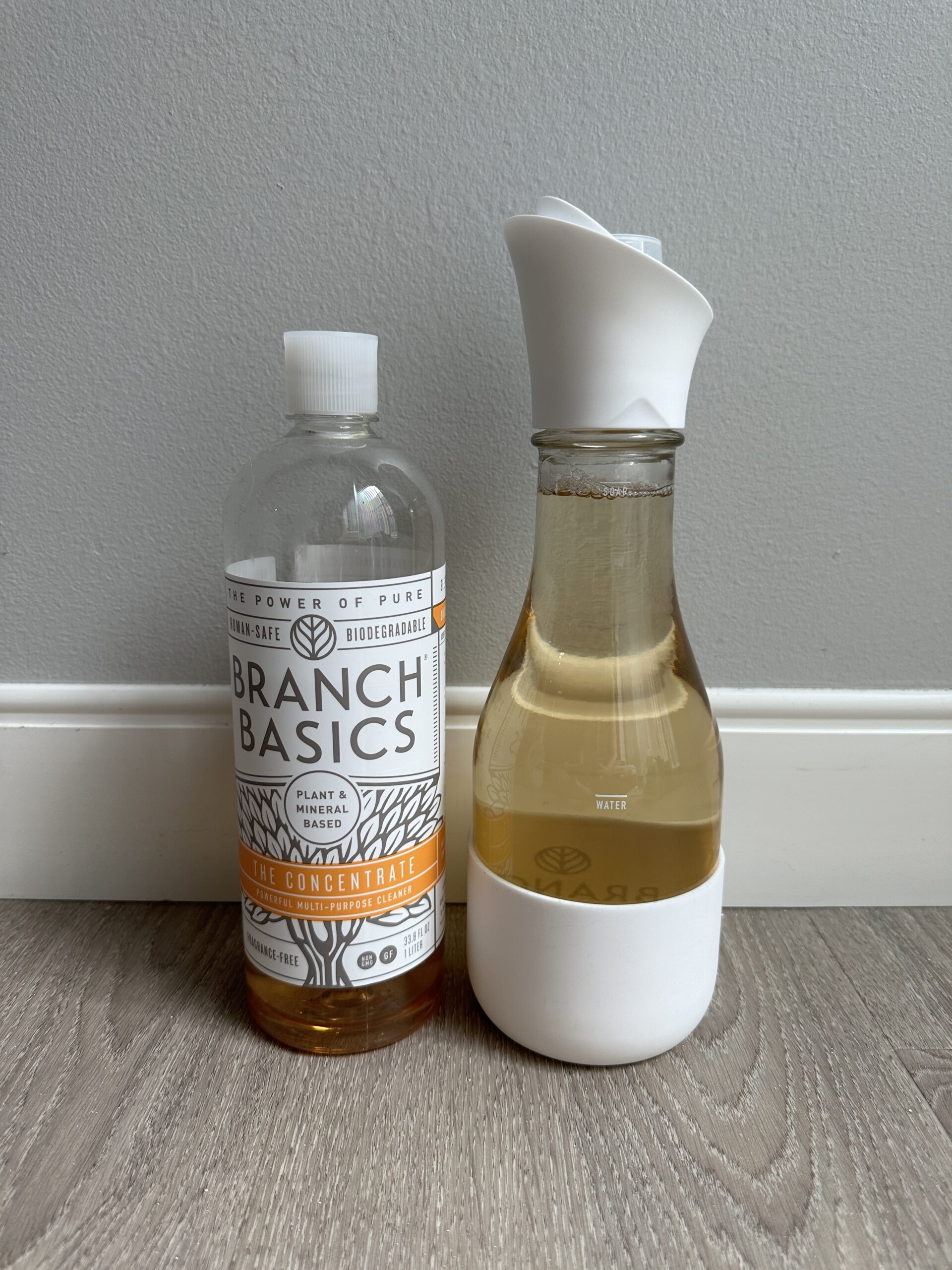 A bottle of Branch Basics concentrate and a glass carafe with a white cap filled partially with a liquid, both placed on a wooden floor against a gray wall.