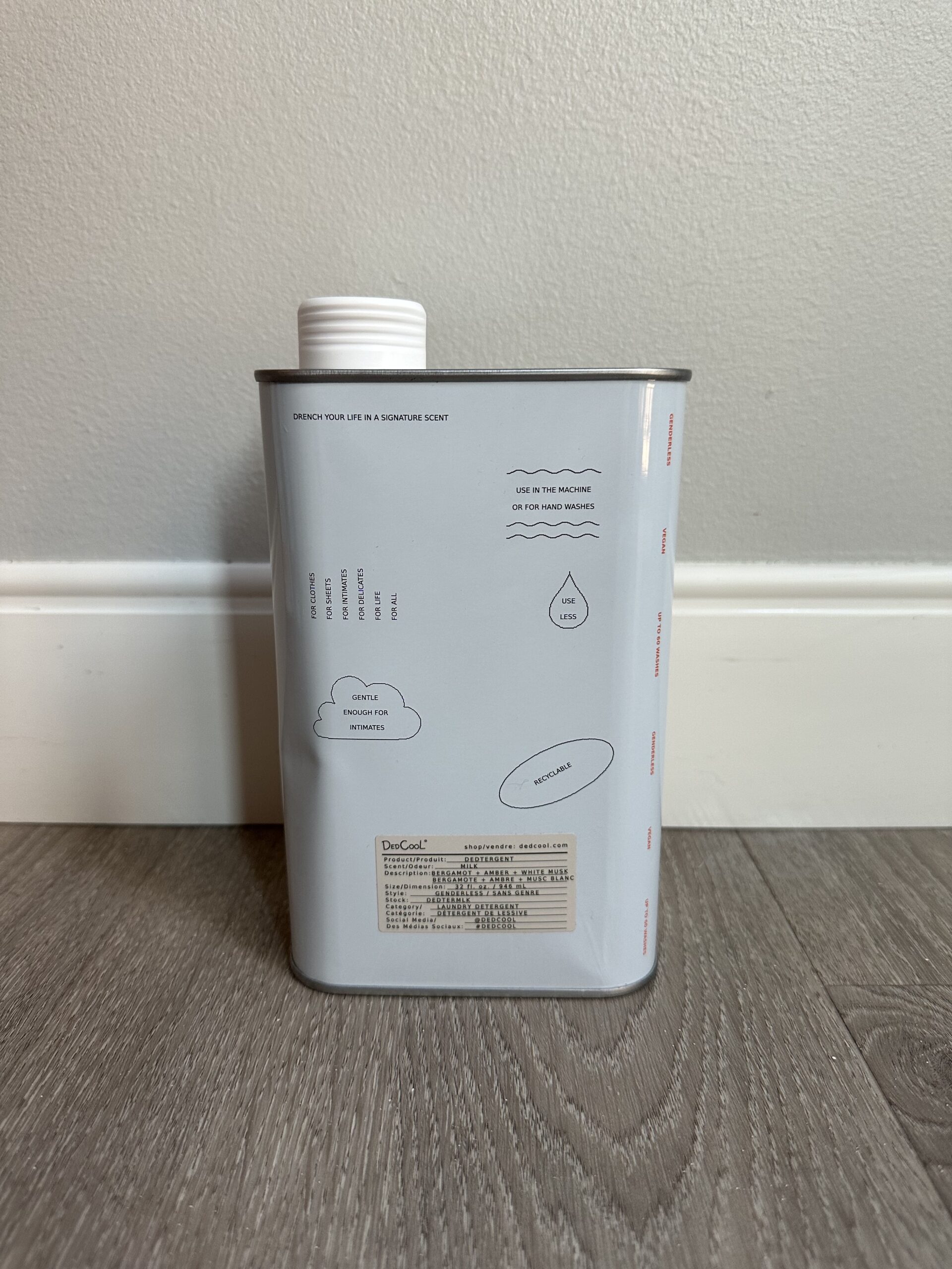 A rectangular gray container with a white cap, standing on a wooden floor against a light gray wall. The container has printed text and minimalistic drawings of mountains, clouds, and waves.