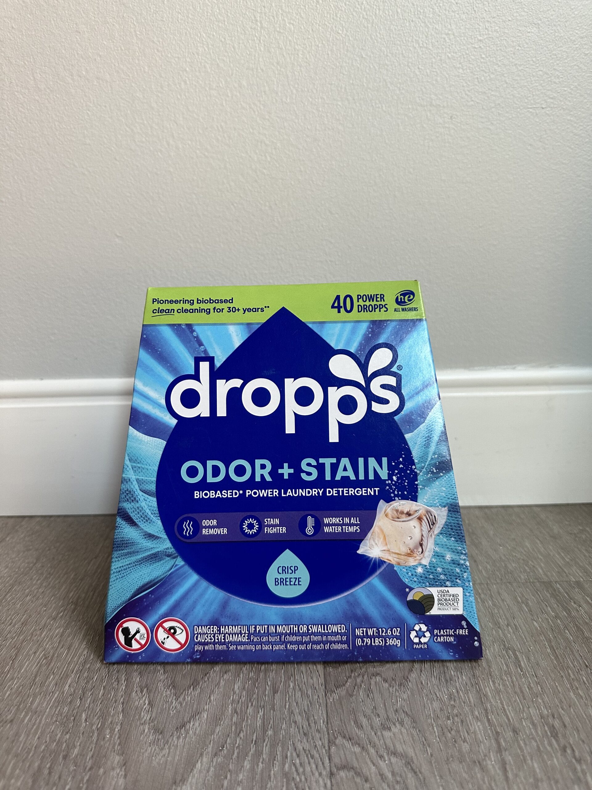 A box of Dropps Odor + Stain Bio-Based Power Laundry Detergent with 40 pods is placed on a wooden floor against a white wall. The box highlights "Crisp Breeze" scent and eco-friendly features.