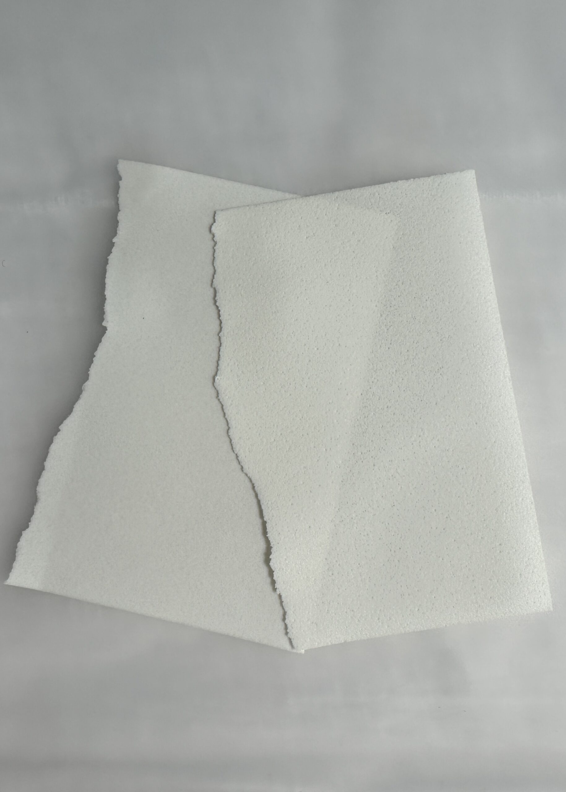 Two sheets of rough-edged white textured paper are stacked unevenly on a gray surface. The top sheet has a torn edge, revealing the texture of the paper.