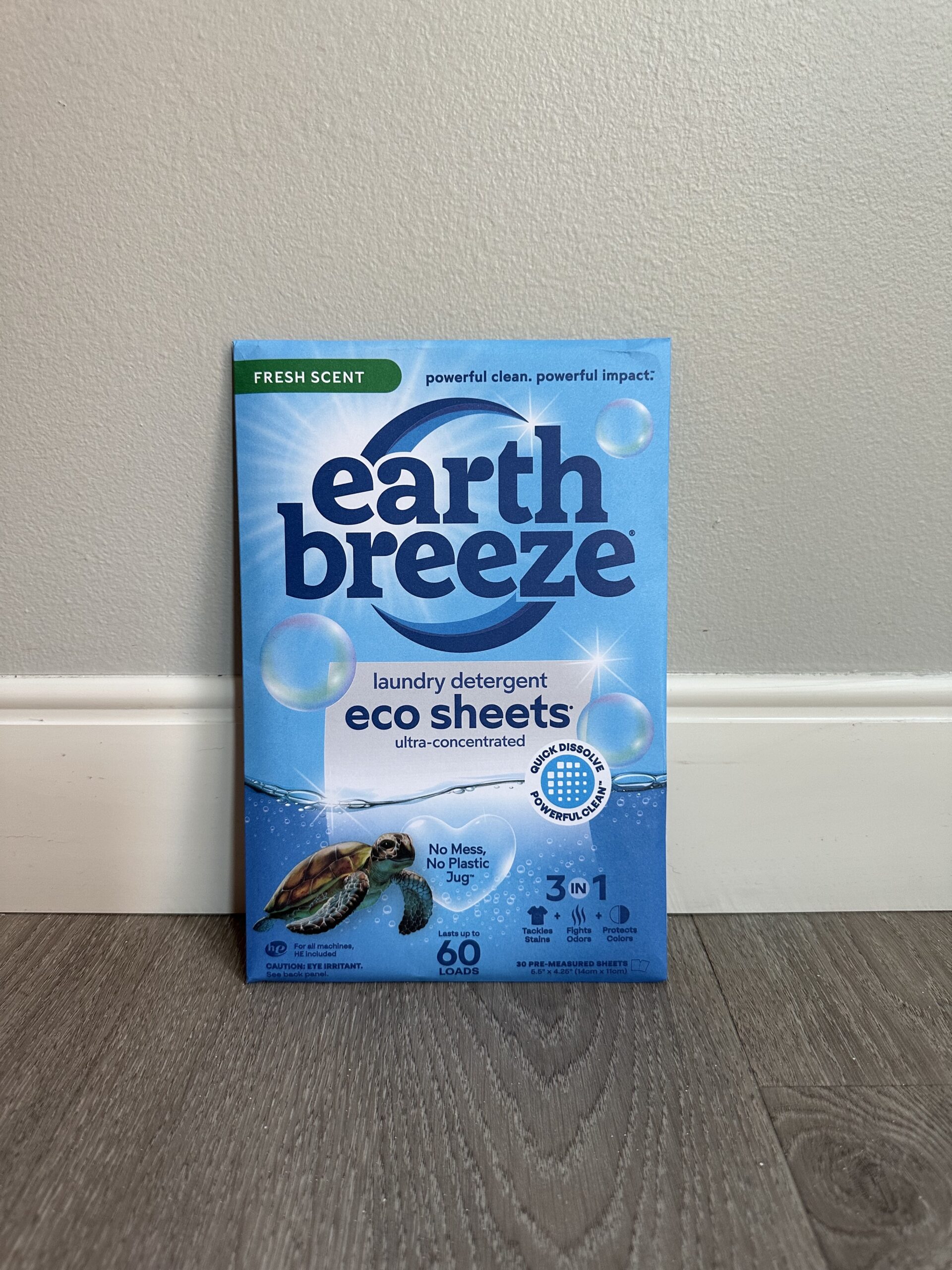 Box of Earth Breeze Fresh Scent eco sheets laundry detergent 3-in-1 placed upright against a grey wall on a wooden floor. The packaging features a turtle and various environmental benefits.