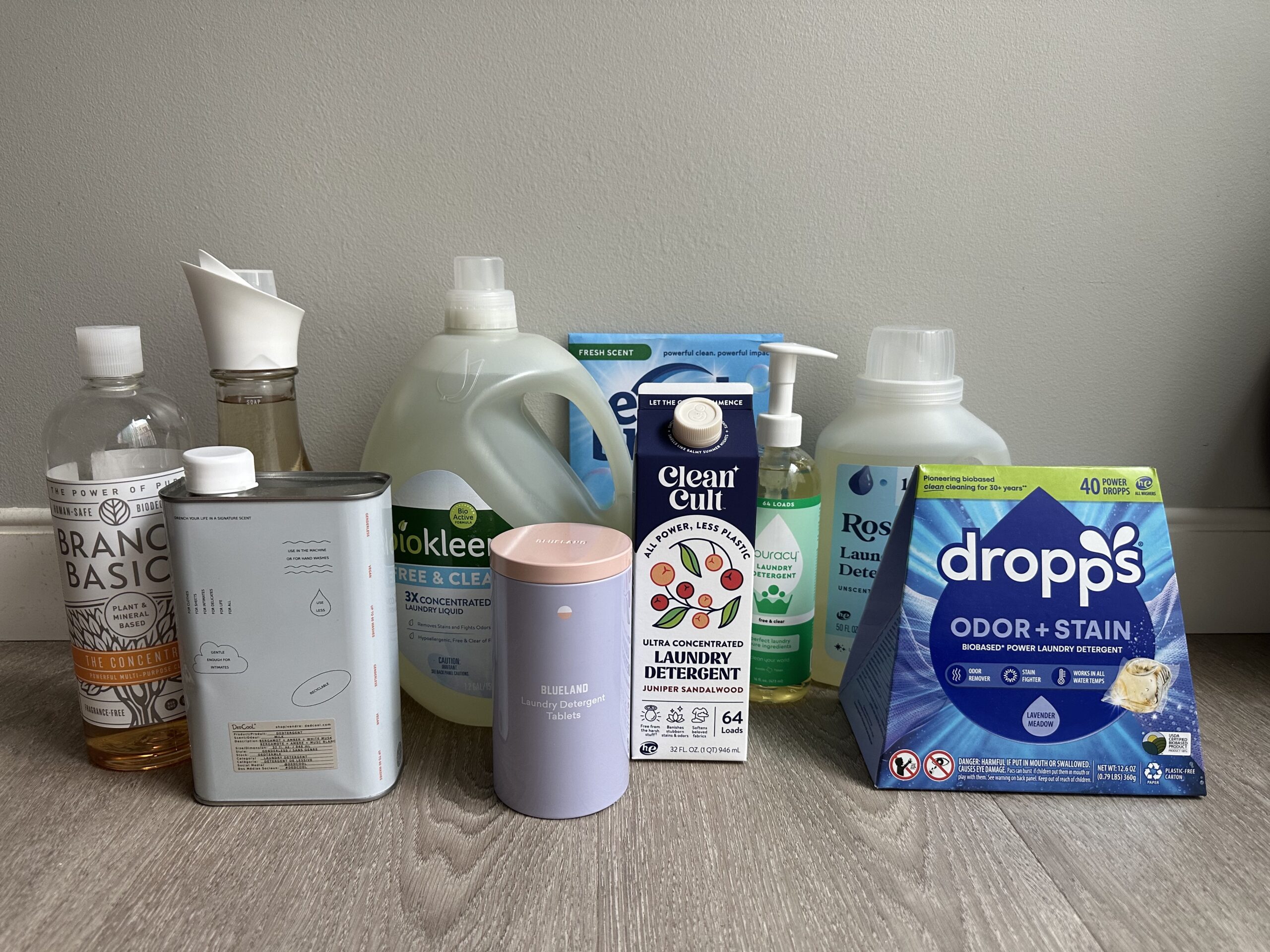 A variety of eco-friendly laundry detergents in different packaging are displayed on a wooden floor against a neutral wall.