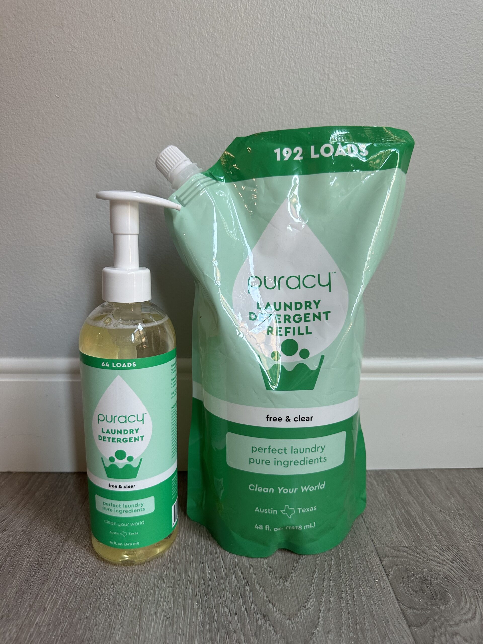 A bottle of Puracy liquid laundry detergent and a refill pouch are placed side by side on a wooden floor. The bottle has a pump dispenser, and both items feature green and white packaging.