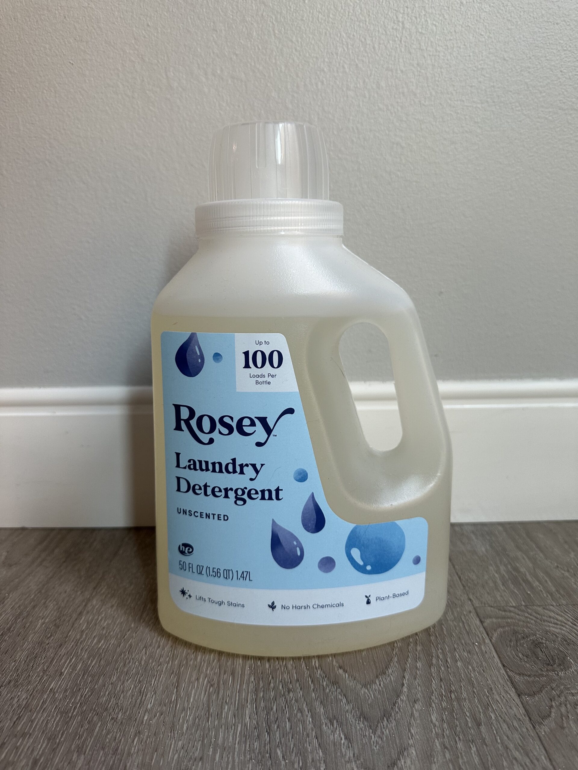 A 50 fl oz bottle of Rosey Unscented Laundry Detergent is placed on a gray wooden floor in front of a light gray wall. The label features blue water droplets and states it is plant-based and chemical-free.