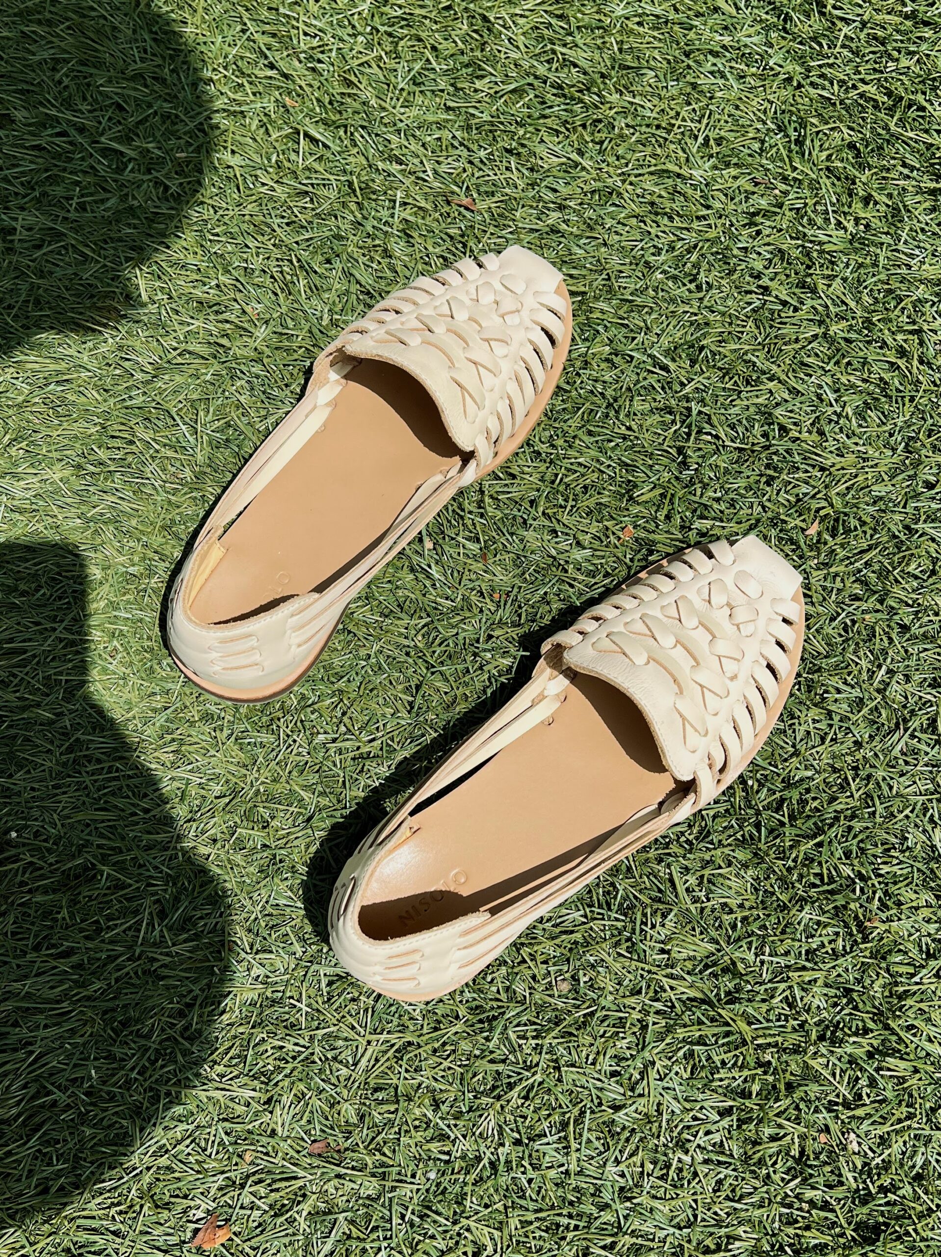 A pair of light-colored woven huarache sandals resting on a grassy surface, with shadows partially visible in the bottom left corner.