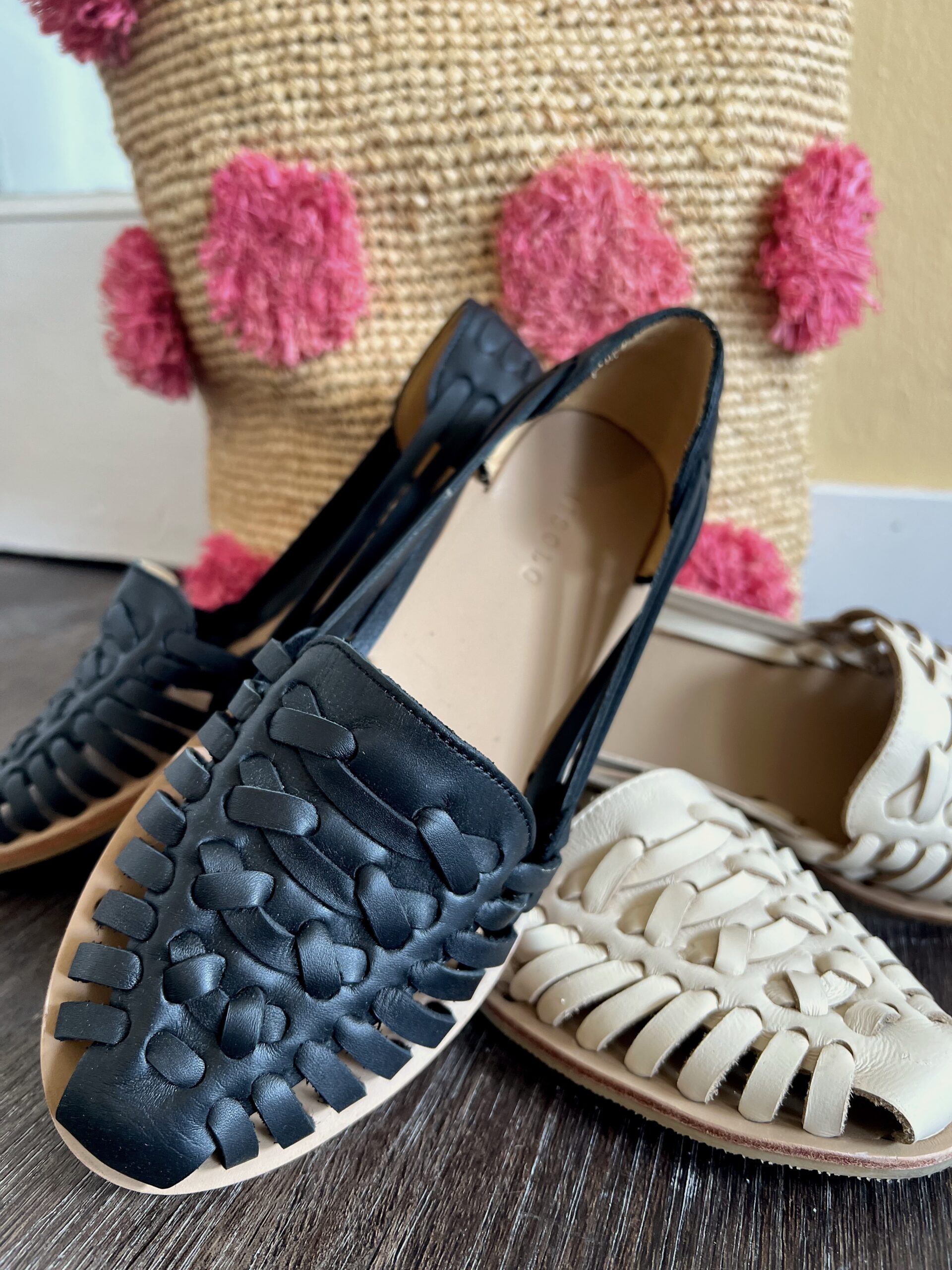 Two pairs of woven shoes, one black and one white, are placed on the floor in front of a woven basket with pink pom-poms. The chic display is reminiscent of traditional huarache sandals, adding a touch of artisanal elegance to the setting.