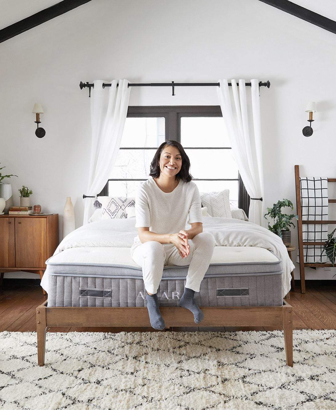 A person sits on a neatly made bed in a bright, modern bedroom with a large window, wooden furniture, and a patterned rug.