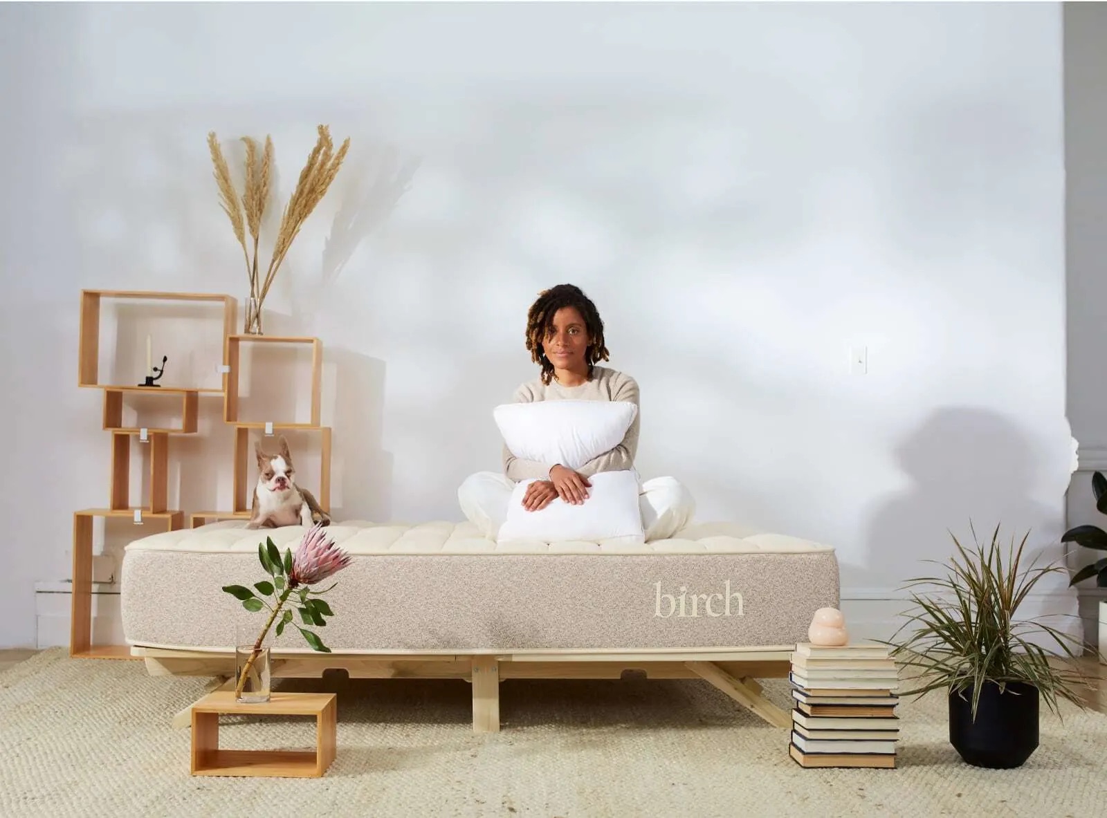 A person sits on a mattress labeled "birch" holding a pillow, with a small dog sitting beside them. The scene includes shelves, a plant, and a neat stack of books.