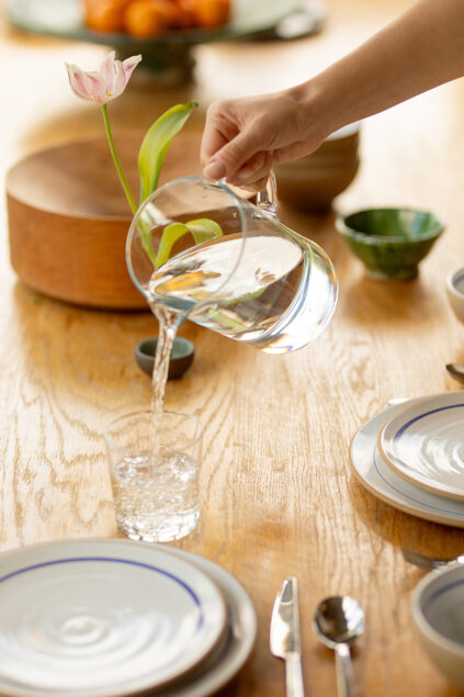 A hand pours water from a glass pitcher into a glass on a wooden table set with plates, bowls, and cutlery, with a pink flower and wooden bowl in the background.