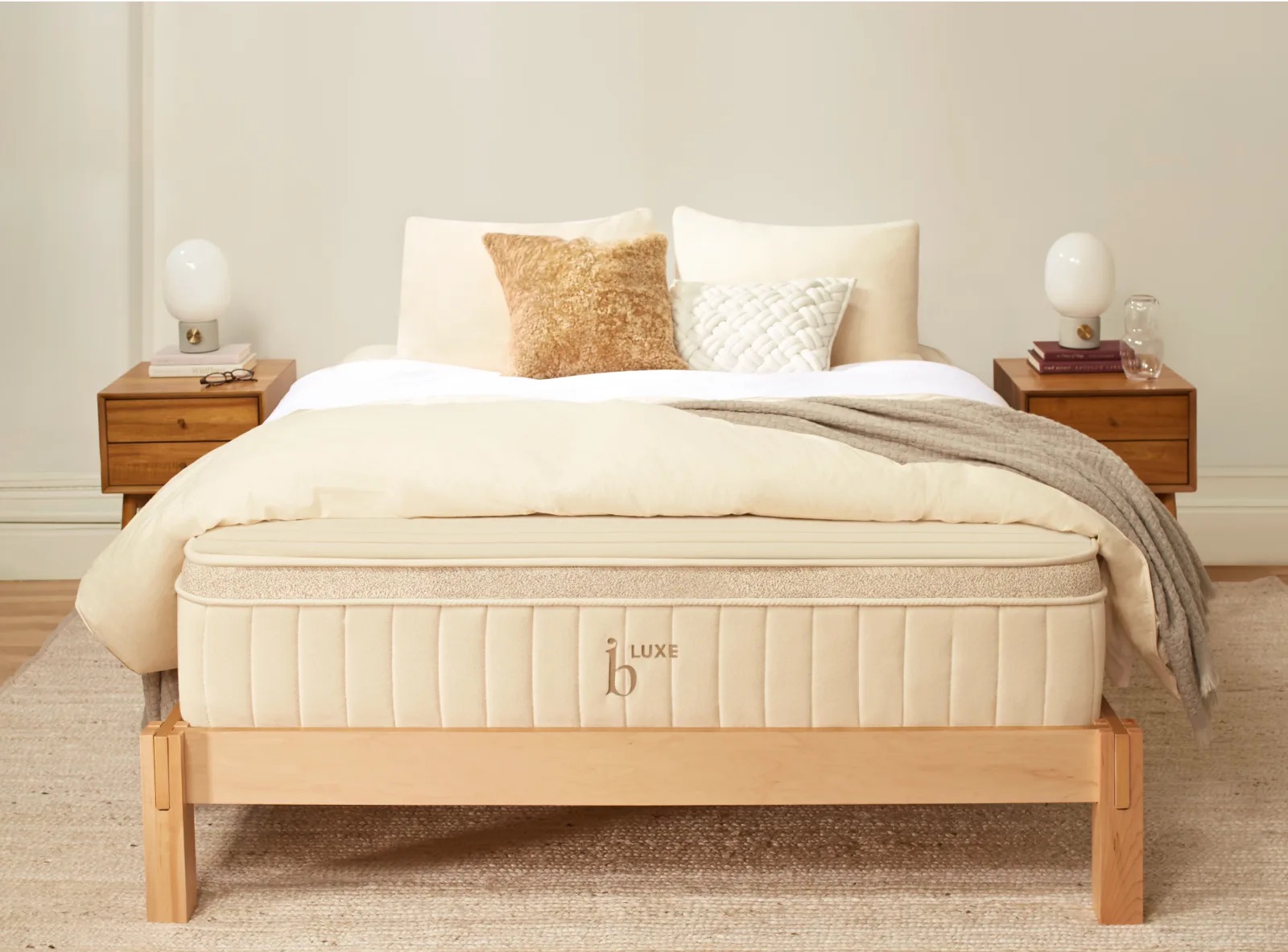 A neatly made bed with creamy white linens, various textured pillows, and a beige mattress labeled "LUXE" on a wooden frame. Two bedside tables with lamps and decor are on each side.