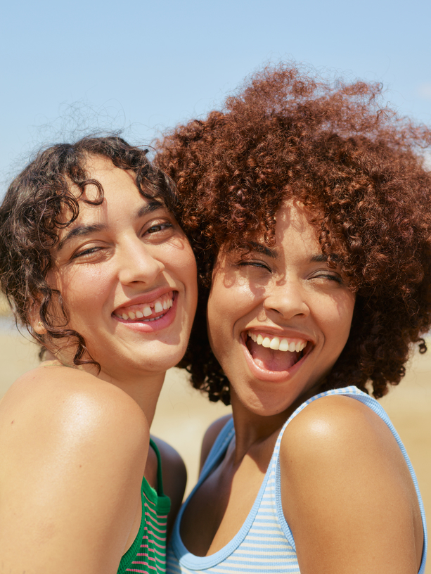 Two people with curly hair smiling and looking at the camera on a sunny day. They are wearing tank tops and appear to be outdoors.