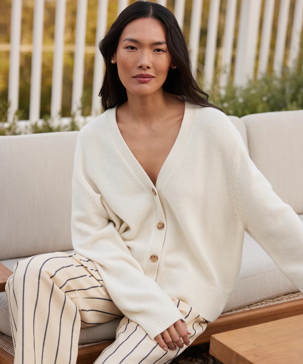 A person with long hair sits on an outdoor couch wearing a white button-up sweater and striped pants.