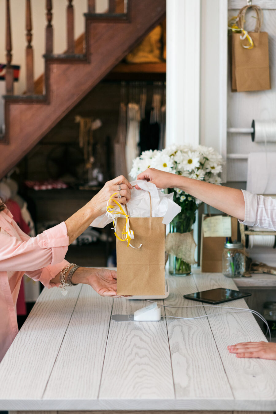 A person hands a brown paper bag with tissue paper to another person across a wooden counter. Flowers and a tablet are on the counter.