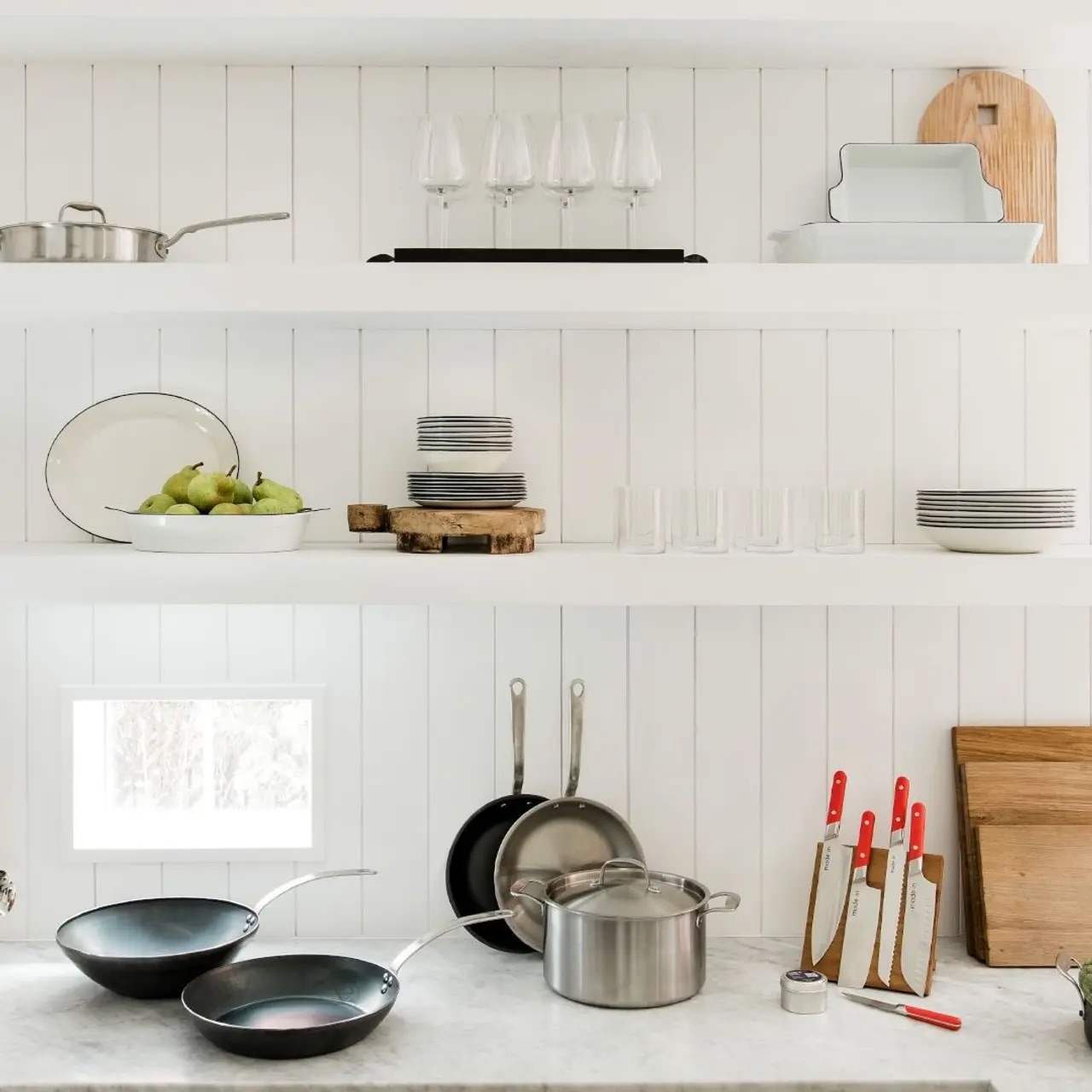 A tidy kitchen shelf with plates, glasses, pears, pots, pans, and knives organized on a white background.