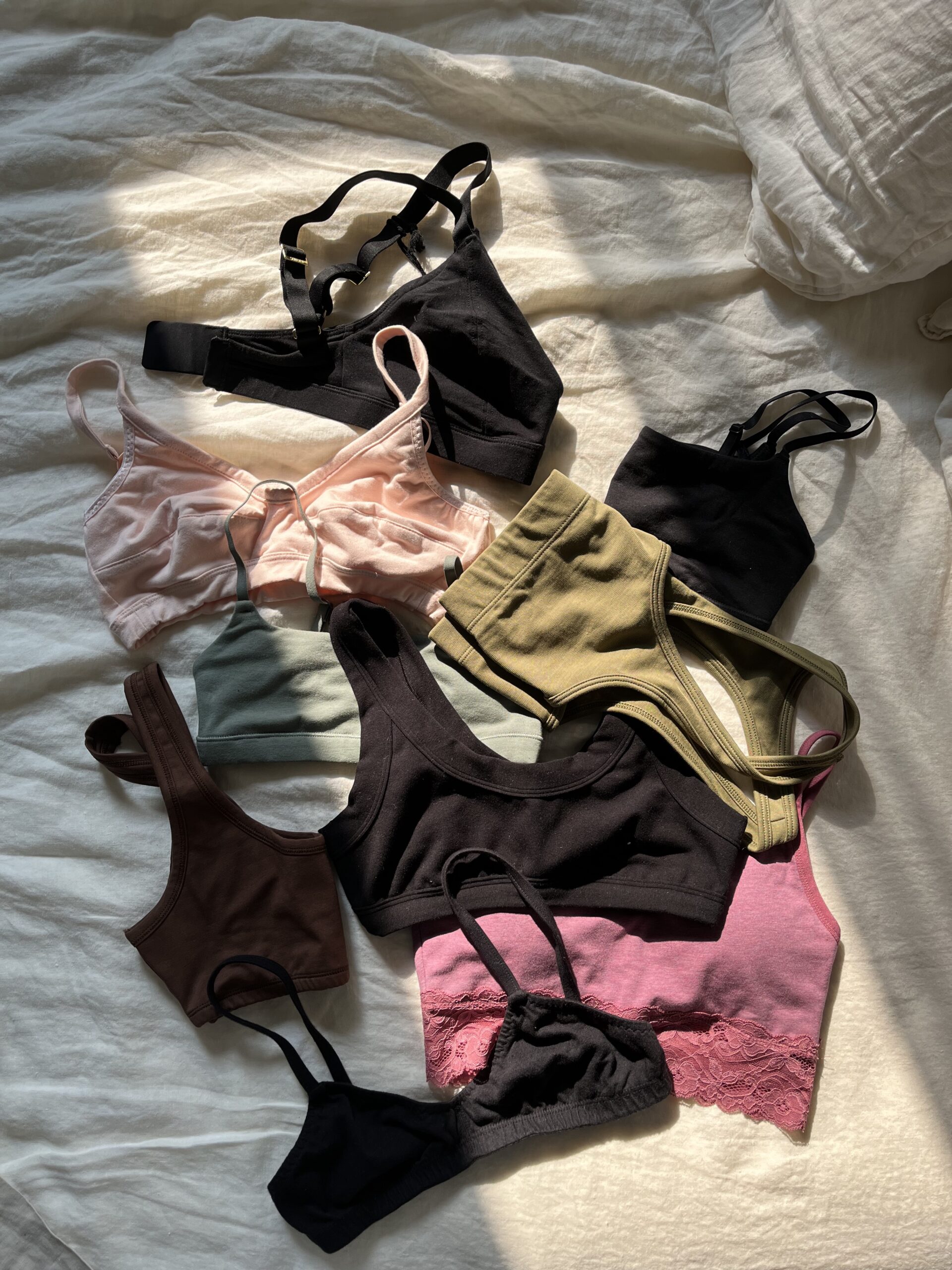 A collection of various colored bras and bralettes laid out on a bed with white sheets, illuminated by natural light.