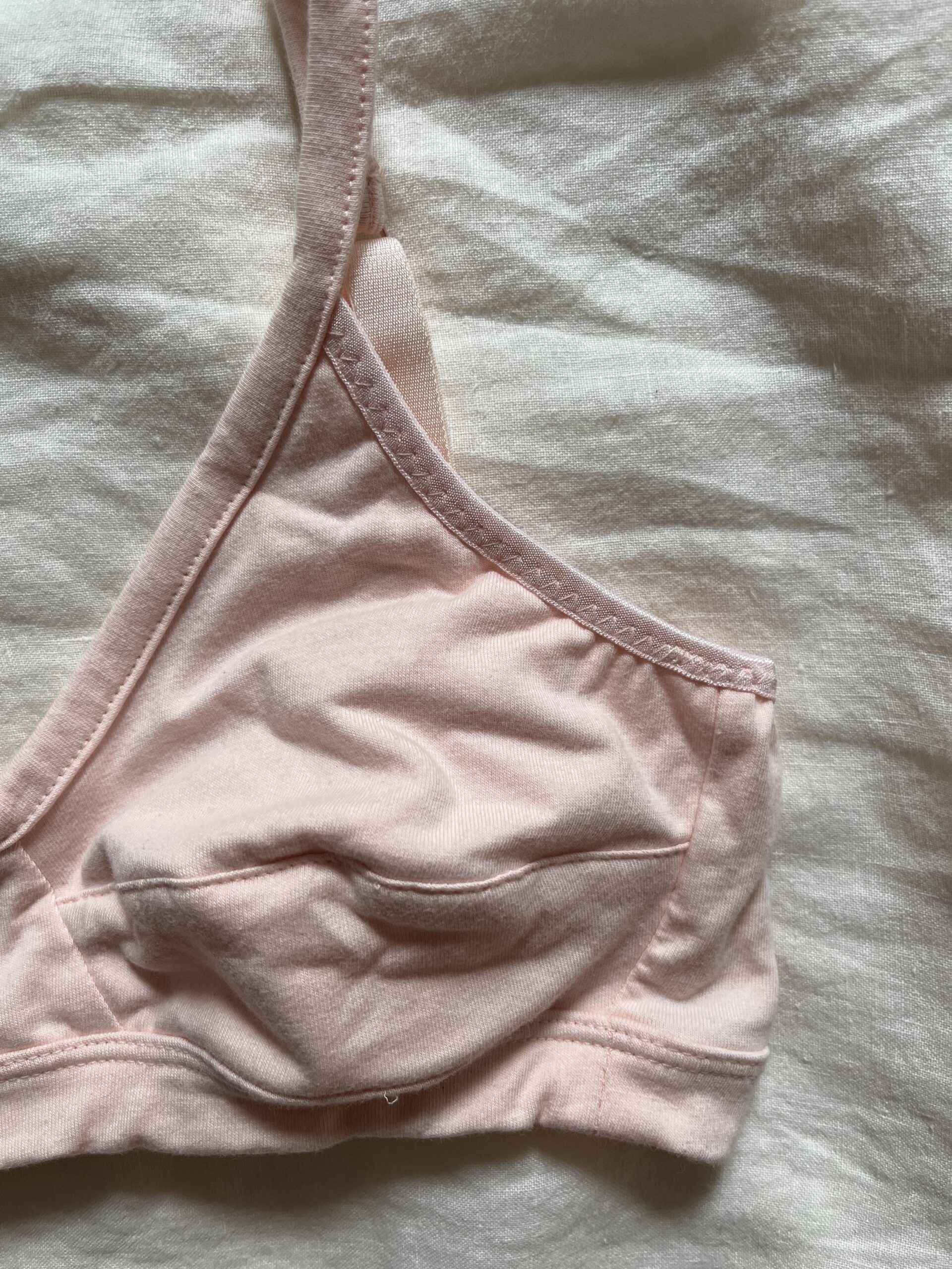 Close-up of a pale pink, unlined bralette with a soft cotton texture, lying flat on a light-colored fabric background.