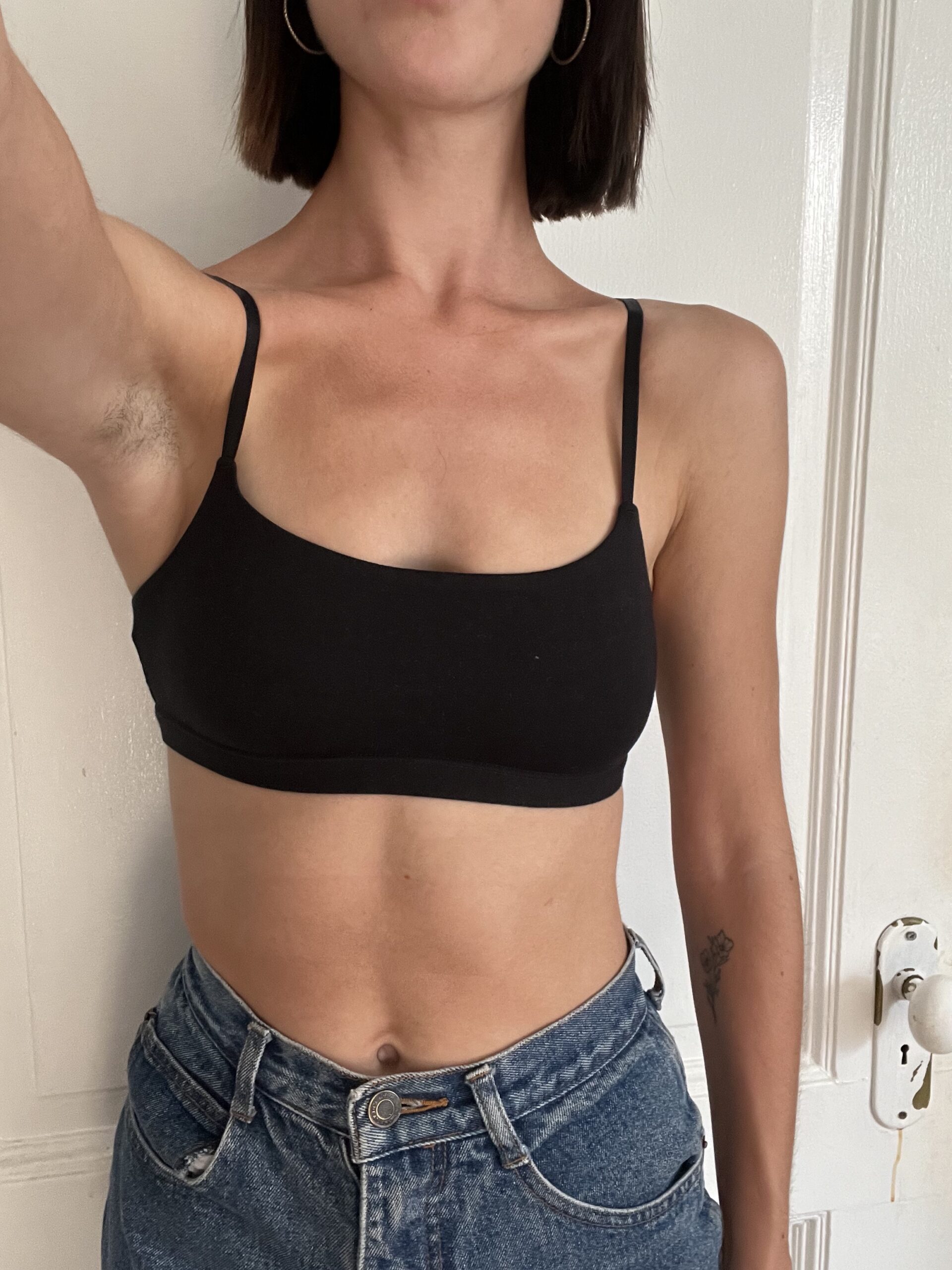 Person wearing a black crop top and blue jeans, standing with an arm raised, in front of a white door.