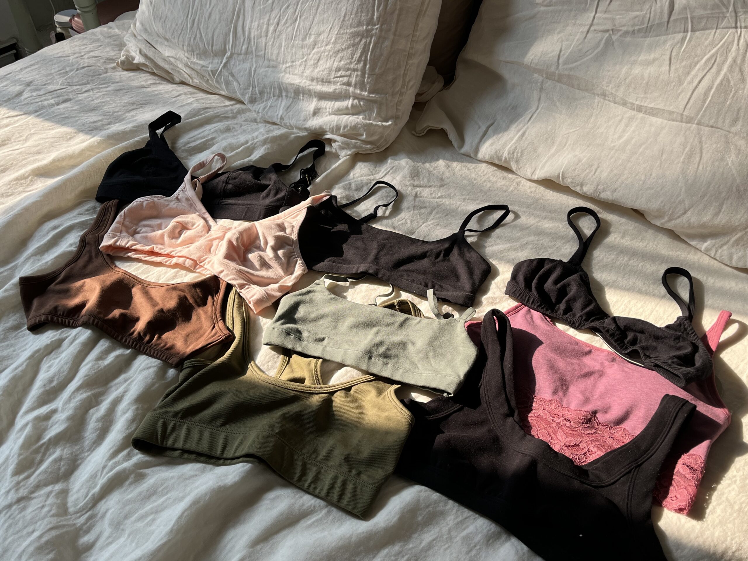 A collection of different colored bras and bralettes are laid out on a bed with white sheets and pillows.