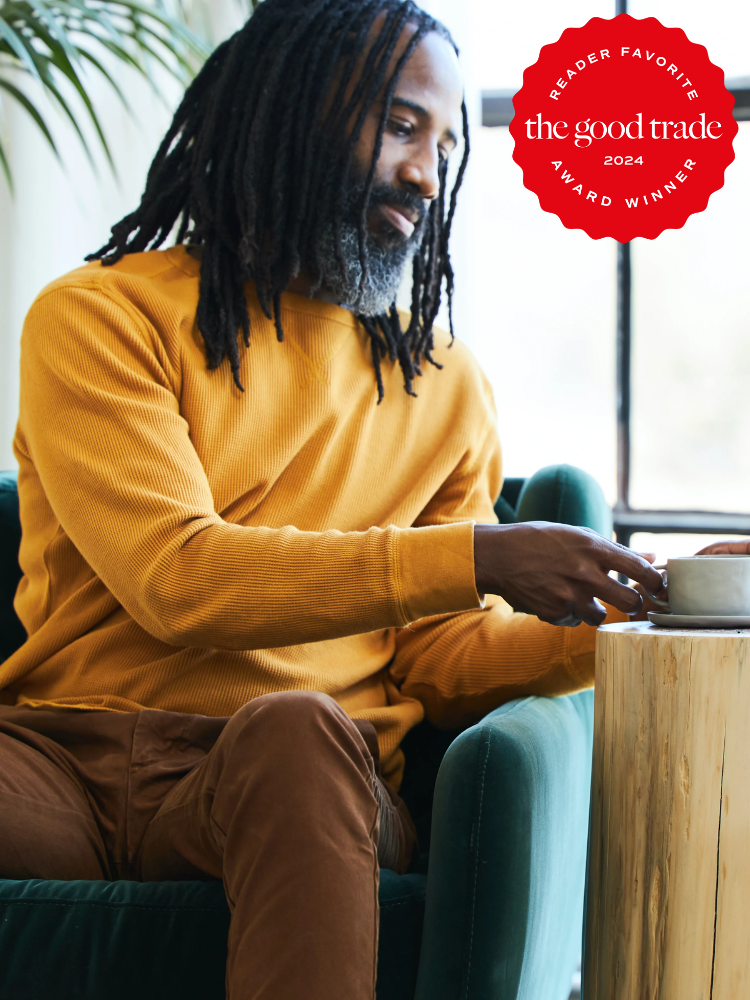 A man with long hair and a beard, wearing a mustard yellow sweatshirt, sits on a green chair and reaches for a cup. A red "Reader Favorite, the good trade, 2024 Award Winner" badge is in the top right corner.