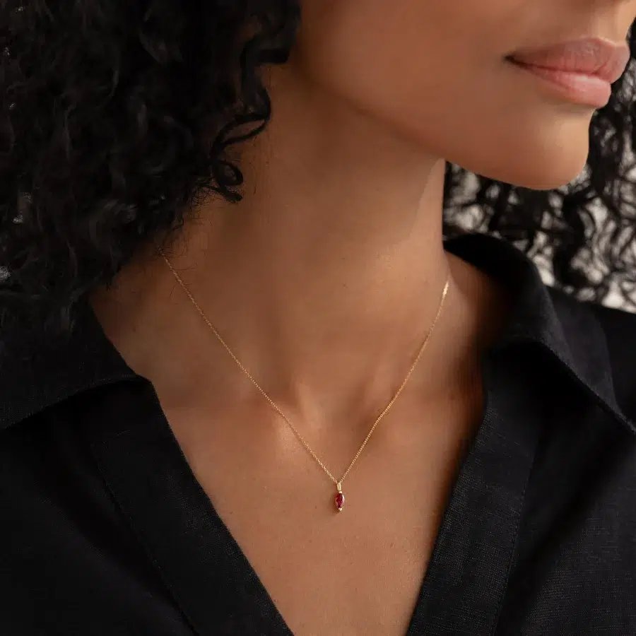 A woman wearing a delicate gold necklace with a small red gemstone pendant. She has curly hair and is dressed in a black shirt.