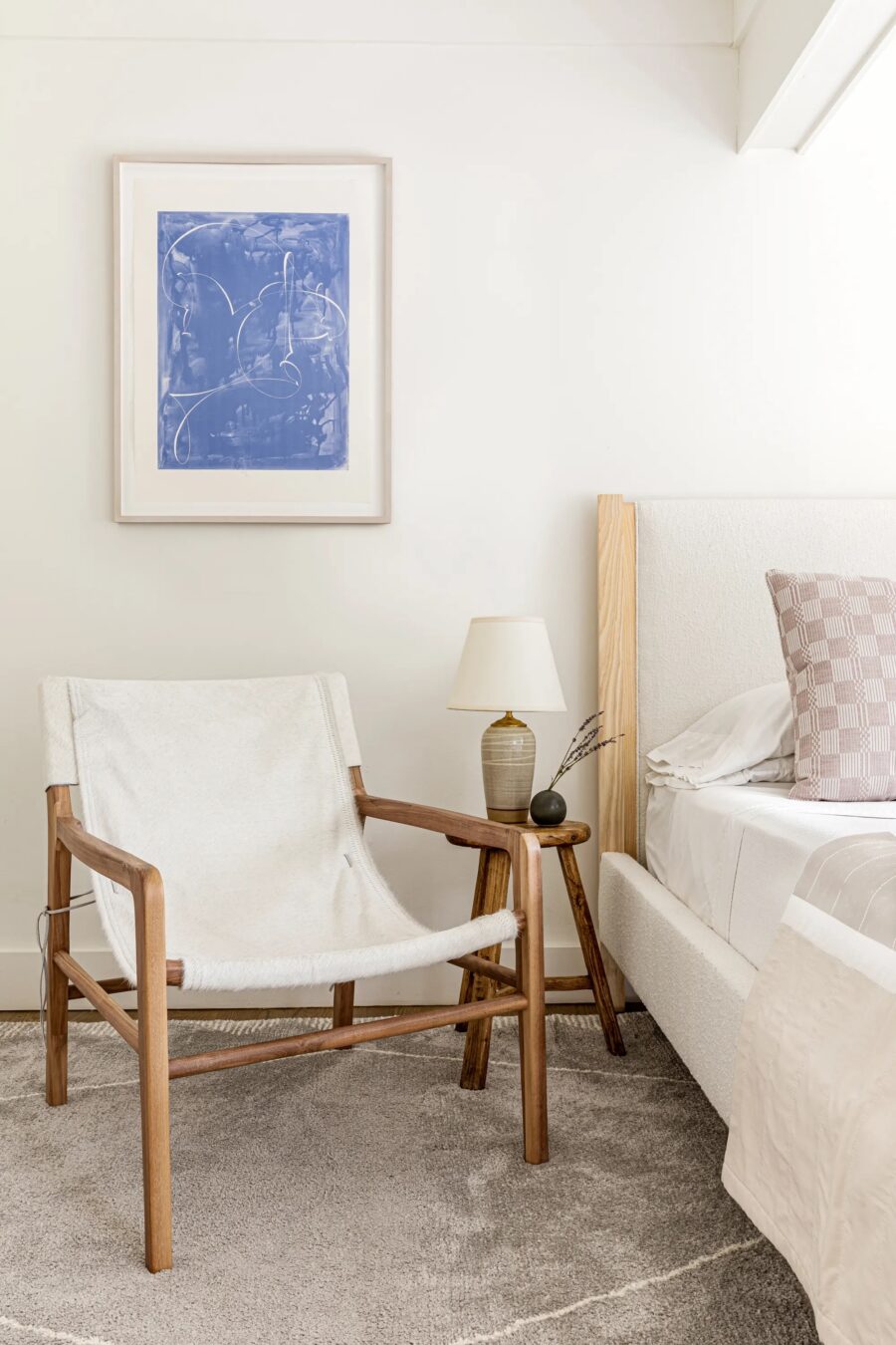 A modern bedroom featuring a wooden chair with a white sling seat, a small wooden side table holding a lamp and decor, and a white bed. A blue abstract painting hangs on the wall above the chair.