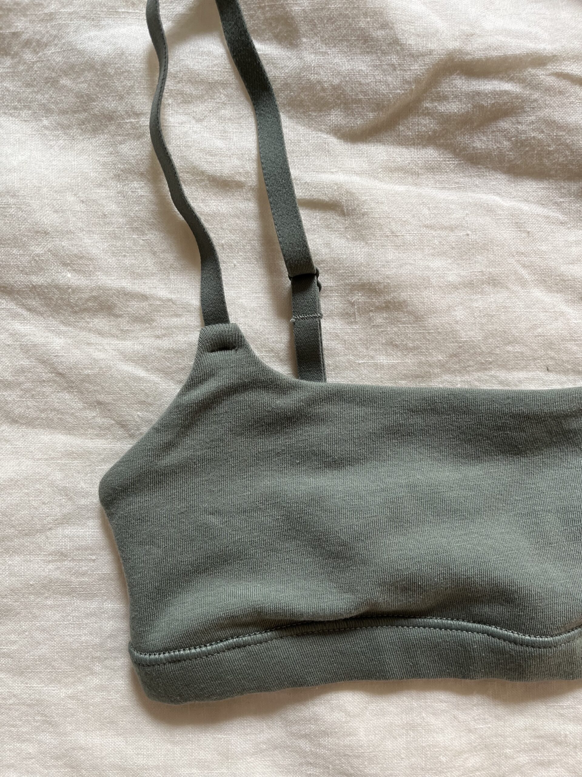 Close-up of a khaki green bralette with thin adjustable straps, lying flat on a light-colored fabric background.