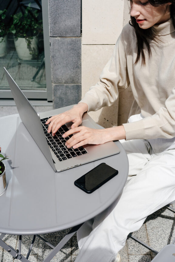 Young female student with brown hair working at cafe table on street using laptop in bright daylight