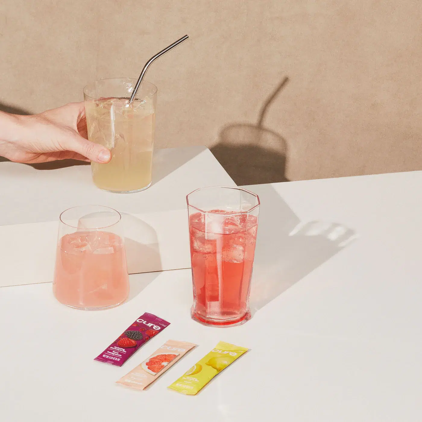 Three glasses filled with different colored beverages are on a white table with three drink mix packets nearby. A hand holds one glass with a metal straw.