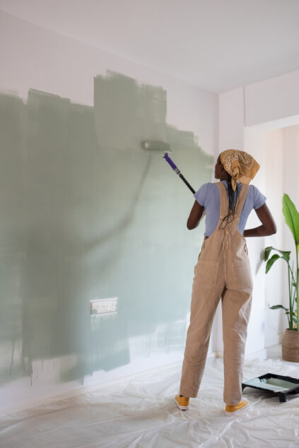 Person painting a white wall green using a roller in a brightly lit room, wearing overalls and a headscarf. Plastic sheet covers the floor below.