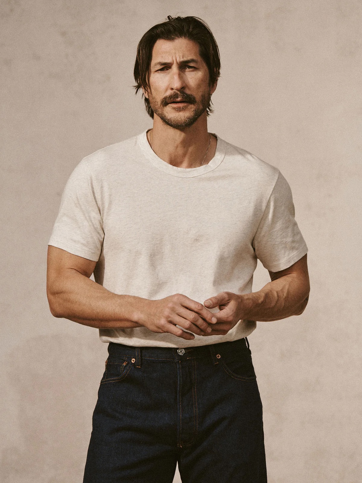 A man with medium-length dark hair and a light beard, wearing a white t-shirt and blue jeans, stands against a neutral background, looking thoughtful.