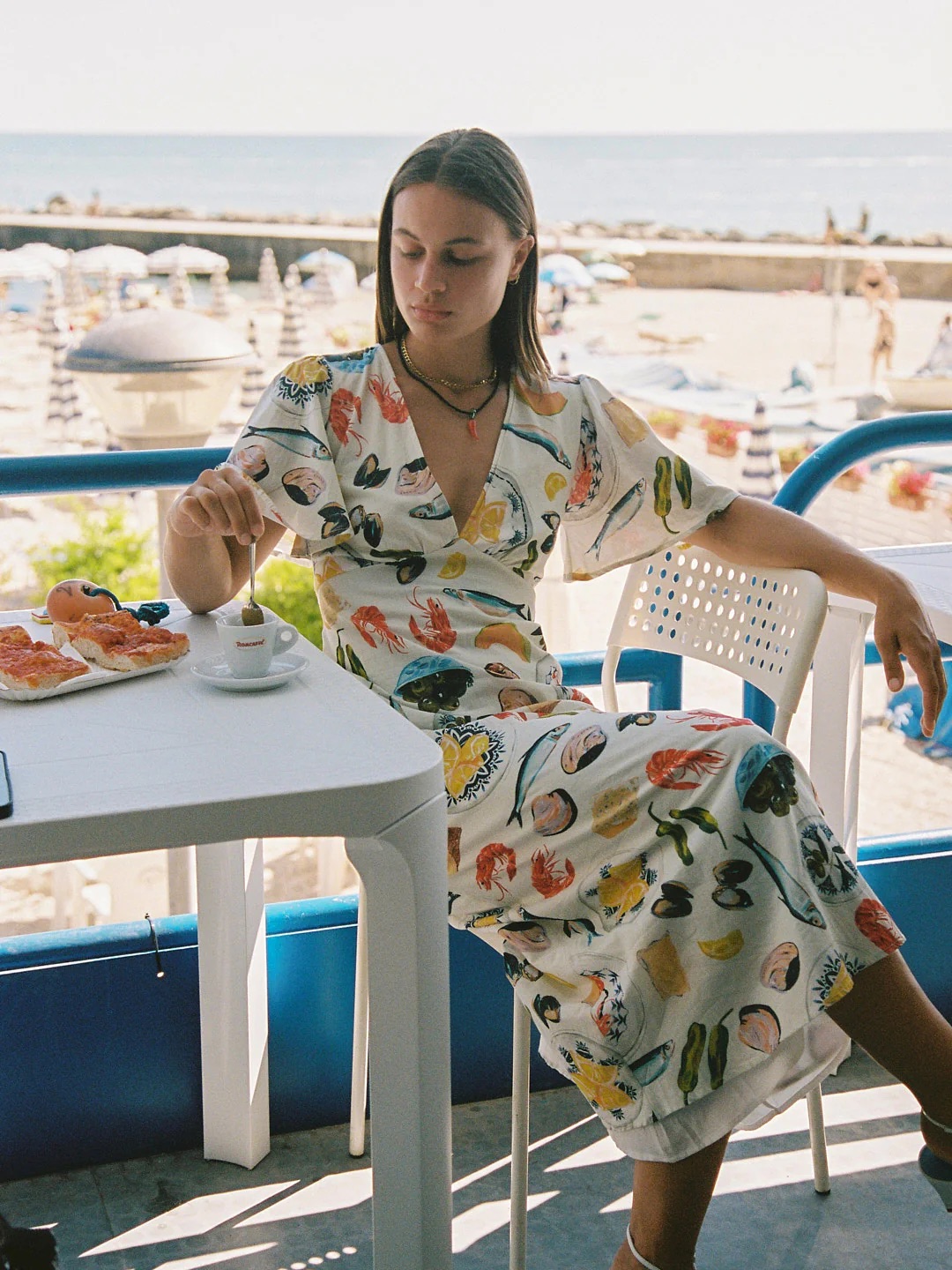 A woman in a colorful dress sits at a seaside café table, stirring her coffee, with boats visible in the background.