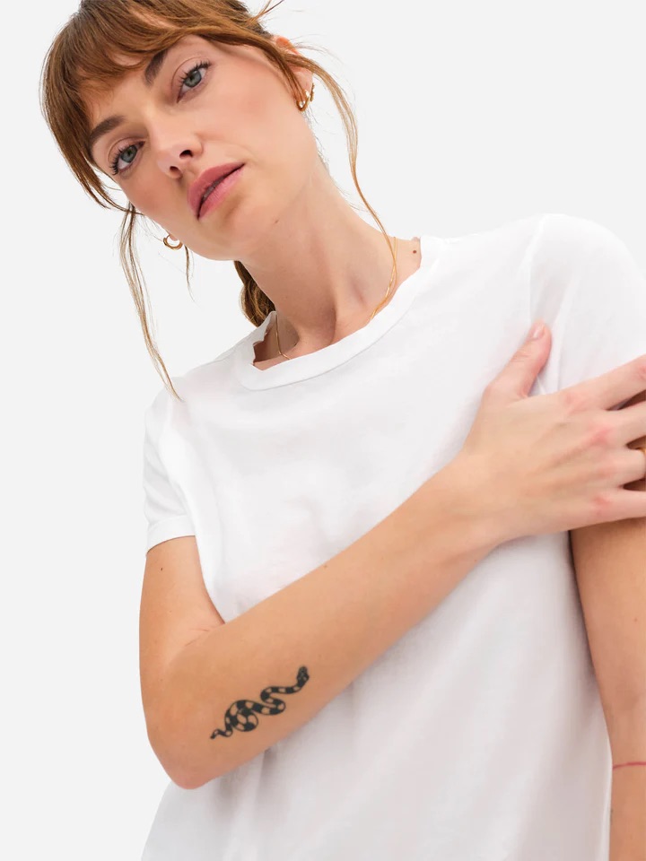 Young woman in a white t-shirt, looking at the camera, with a small black tattoo on her left arm.