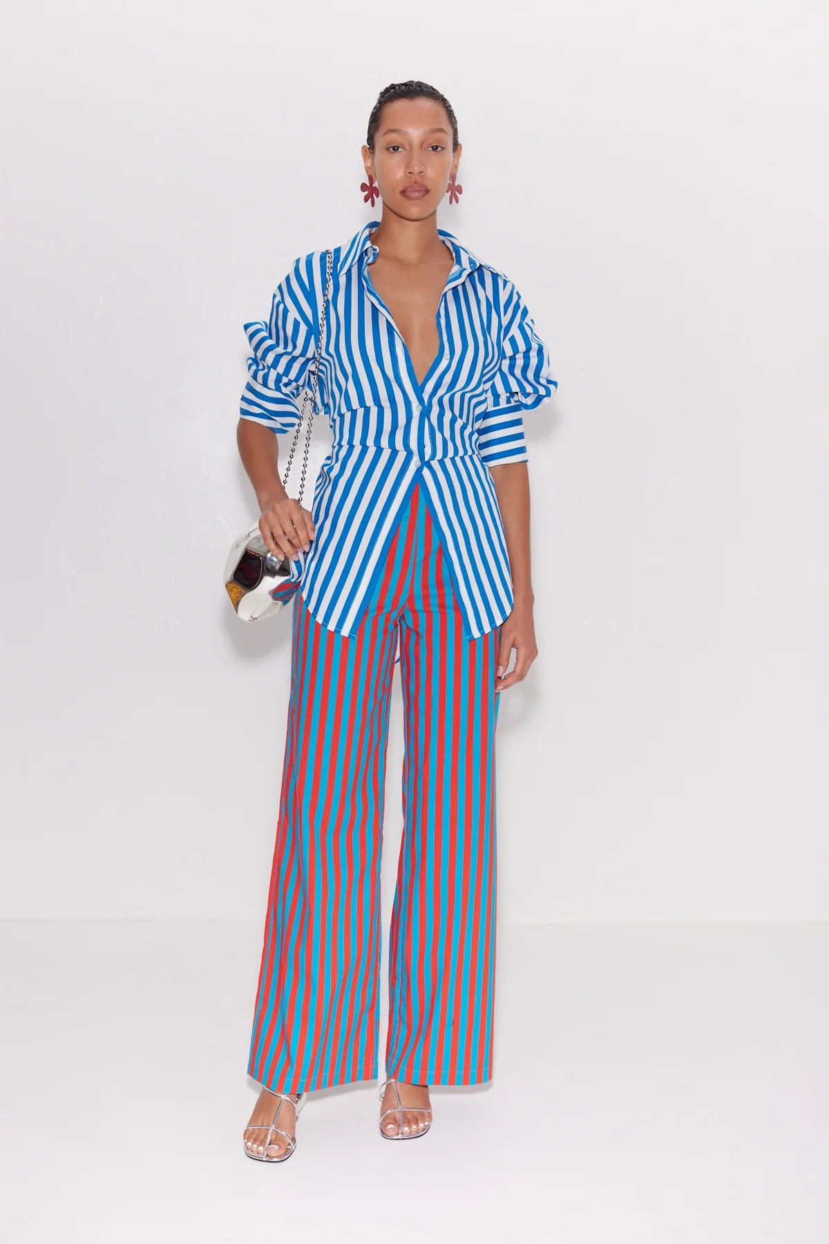 A woman wearing a bold striped jumpsuit with ruffled sleeves, holding a metallic purse, standing against a plain white background.